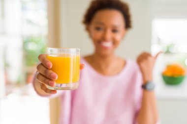 Genç afro-amerikan kadın evde işaret ve mutlu yüzü gülümseyerek yan başparmak ile gösteren portakal suyu driking