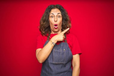 Kırmızı izole arka plan üzerinde önlük üniforma giyen orta yaş yaşlı kadın yan parmak ile işaret sürpriz, açık ağız hayret ifade.