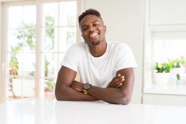 Beyaz masada yakışıklı Afrikalı Amerikalı adam evde mutlu yüz kamera bakarak çapraz silah ile gülümsüyor. Pozitif kişi.