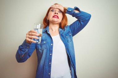 Genç güzel kızıl saçlı kadın beyaz izole arka plan üzerinde su cam içme baş üzerinde el ile vurguladı, utanç ve sürpriz yüz ile şok, kızgın ve sinirli. Korku ve hata için üzgün.