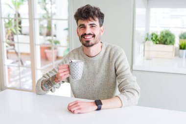 Genç adam sabah ları mutlu bir yüzle bir fincan kahve içerken ayakta ve kendinden emin bir gülümsemeyle gülümseyerek dişlerini gösteriyor.