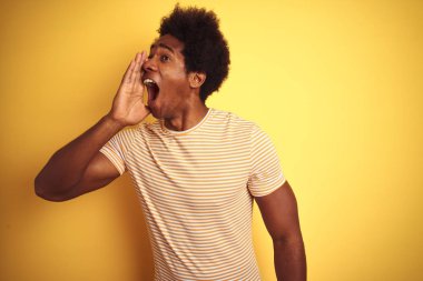 Afro saçlı Amerikalı adam çizgili t-shirt giyen izole sarı arka plan üzerinde bağırarak ve ağız da el ile yan yüksek sesle bağırıyor. İletişim kavramı.