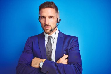 Genç yakışıklı operatör adam mavi izole arka plan şüpheci ve sinir üzerinde çağrı merkezi kulaklık giyen, çapraz kolları ile yüzünde onaylamayan ifade. Negatif kişi.