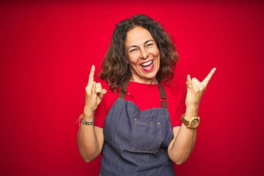 Orta yaş yaşlı kadın kırmızı izole arka plan üzerinde önlük üniforma giyen deli ifade elleri ile rock sembolü yapıyor bağırarak. Müzik yıldızı. Ağır müzik konsepti.