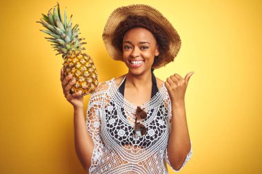 Sarı izole arka plan üzerinde pineaple tutan afro saçlı genç afro-amerikan kadın işaret ve mutlu yüz gülümseyerek yan başparmak ile gösteren