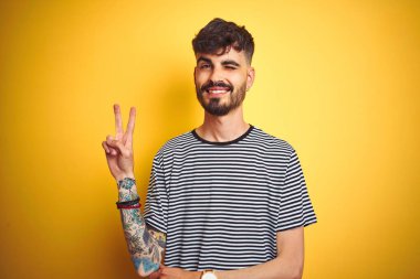 Çizgili t-shirt giyen dövme giyen genç adam mutlu yüz zafer işareti yapıyor kameraya göz kırparak ile gülümseyerek izole sarı arka plan üzerinde duruyor. İki numara.