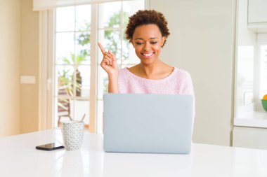 Genç afro-amerikan kadın yüzünde büyük bir gülümseme ile bilgisayar dizüstü bilgisayar kullanarak çalışan, kameraya bakarak yan el ve parmak ile işaret.