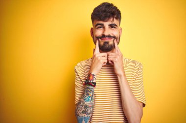 Çizgili t-shirt giyen dövme giyen genç adam izole sarı arka plan üzerinde duran açık ağız ile gülümseyerek, parmakları işaret ve neşeli gülümseme zorluyor