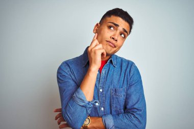 Denim gömlek giyen genç brezilyalı adam elini çeneüzerinde sorgularken izole beyaz arka plan üzerinde durarak, dalgın ifade. Düşünceli bir yüzle gülümsüyor. Şüphe kavramı.