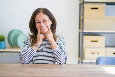 Orta yaş yaşlı kadın evde masada oturan açık ağız ile gülümseyen, parmakları işaret ve neşeli gülümseme zorlama
