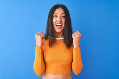 Turuncu tişört giyen genç güzel Çinli kadın izole mavi arka plan üzerinde ayakta kutluyor şaşırmış ve kollarını kaldırdı ve gözleri açık başarı için şaşırttı. Kazanan kavramı.
