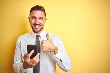 Genç yakışıklı iş adamı sarı izole arka plan üzerinde akıllı telefon kullanarak büyük bir gülümseme ok işareti yapıyor mutlu, parmakları ile başparmak, mükemmel işareti