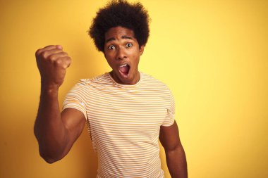 Afro saçlı amerikalı adam çizgili t-shirt izole sarı arka plan üzerinde duran giyen kızgın ve öfke ile bağırırken öfkeli ve öfkeli yumruk yükselterek. Öfke ve agresif kavram.