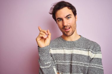 Genç yakışıklı adam izole pembe arka plan üzerinde duran rahat çizgili kazak giyen çok mutlu yan el ve parmak ile işaret