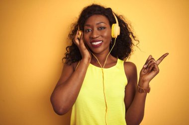 Izole sarı arka plan üzerinde kulaklık kullanarak müzik dinleyen Afrikalı Amerikalı kadın çok mutlu yan el ve parmak ile işaret