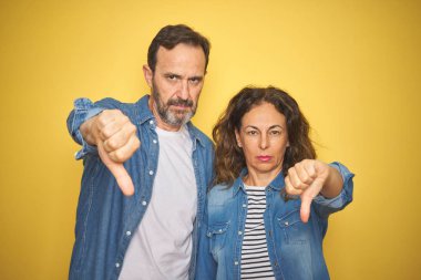 Güzel orta yaş çift birlikte izole sarı arka plan üzerinde kot gömlek giyen mutsuz ve kızgın gösteren reddi gösteren ve aşağı jest başparmak ile negatif. Kötü ifade.