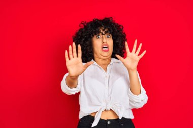 Kıvırcık saçlı genç arap kadın izole kırmızı arka plan üzerinde beyaz rahat gömlek giyen korkmuş ve korku ifade ile korkmuş elleriyle jest durdurmak, şok içinde bağırarak. Panik kavramı.