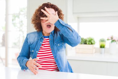 Evde kot ceket giyen kıvırcık saçlı orta yaş yaşlı kadın şok yüz ve gözleri el ile kaplayan gözetleme, utanmış ifade ile parmaklarından bakarak.