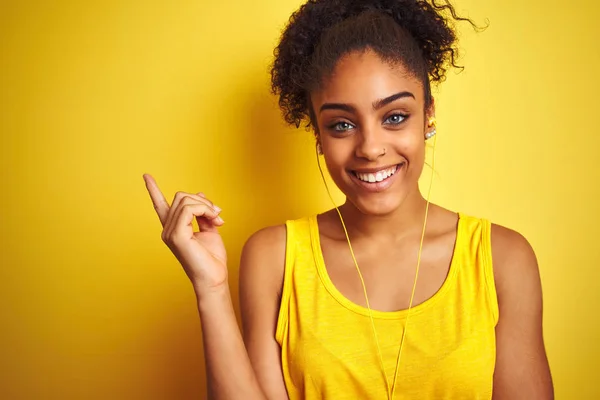 African american woman listening to music using earphones over isolated yellow background with a big smile on face, pointing with hand and finger to the side looking at the camera.