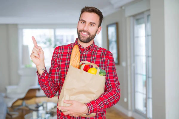 Handsome man holding groceries bag very happy pointing with hand and finger to the side