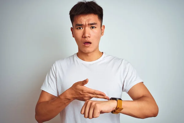 Young asian chinese man wearing t-shirt standing over isolated white background In hurry pointing to watch time, impatience, upset and angry for deadline delay