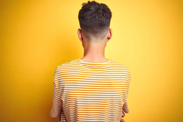 孤立した黄色の背景の上に立つストライプのTシャツを着たタトゥーをした若い男は 十字架の腕で後ろ向きに立っている — ストック写真