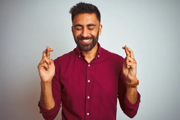 Young indian man wearing red elegant shirt standing over isolated grey background gesturing finger crossed smiling with hope and eyes closed. Luck and superstitious concept.