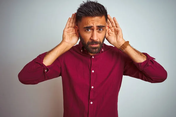 Young indian man wearing red elegant shirt standing over isolated grey background Trying to hear both hands on ear gesture, curious for gossip. Hearing problem, deaf