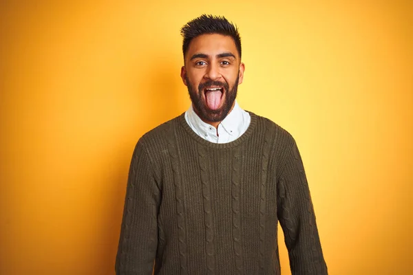 Young indian man wearing green sweater and shirt standing over isolated yellow background sticking tongue out happy with funny expression. Emotion concept.