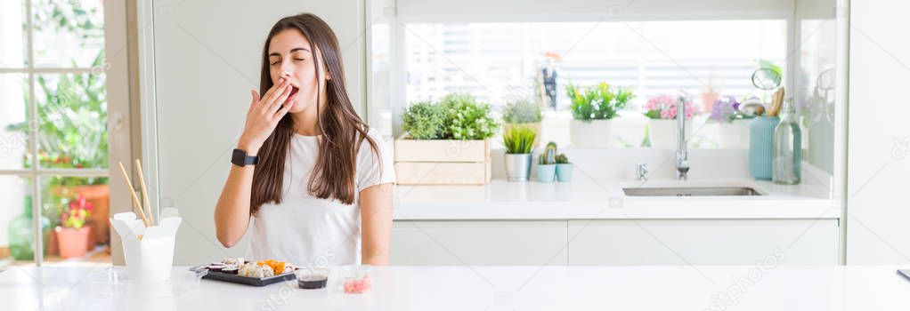 Wide angle picture of beautiful young woman eating asian sushi from delivery bored yawning tired covering mouth with hand. Restless and sleepiness.
