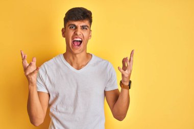 Beyaz tişört giyen genç hintli adam izole sarı arka plan üzerinde duran deli ve deli bağırarak ve agresif ifade ve kollar kaldırdı ile bağırıyor. Hayal kırıklığı kavramı.