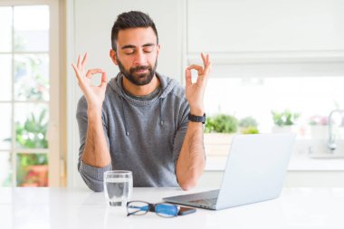 Yakışıklı İspanyol adam bilgisayar dizüstü bilgisayar kullanarak çalışan dinlenmek ve gözleri parmakları ile meditasyon jest yapıyor kapalı gülümseyerek. Yoga konsepti.