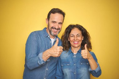 Güzel orta yaş çift birlikte izole sarı arka plan üzerinde el ile jest kadar mutlu başparmak yapıyor ayakta. Başarı gösteren kameraya bakarak ifadeyi onaylama.