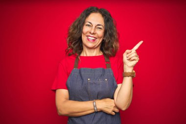 Yüzünde büyük bir gülümseme ile kırmızı izole arka plan üzerinde önlük üniforma giyen orta yaş yaşlı kadın, kameraya bakarak yan el parmağı ile işaret.