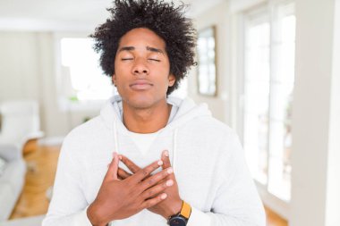 Afro-Amerikan adam kapalı gözleri ve yüzünde minnettar jest ile göğüs üzerinde gülümseyen sweatshirt giyiyor. Sağlık kavramı.