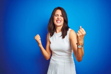 Mavi izole arka plan üzerinde duran beyaz zarif elbise giyen genç iş kadını çok mutlu ve heyecanlı kolları kaldırdı ile kazanan jest yapıyor, gülümseyerek ve başarı için çığlık. Kutlama konsepti.