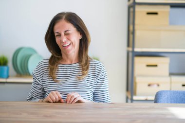 Orta yaş yaşlı kadın evde masada oturan seksi ifade, neşeli ve mutlu yüz ile kameraya bakarak göz kırpıyor.