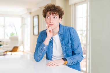 Çene düşünme hakkında soru, dalgın ifade üzerine el ile evde rahat denim ceket giyen genç yakışıklı adam. Düşünceli yüzüyle gülümsüyor. Şüphe kavramı.