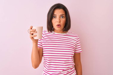 Genç güzel kadın izole pembe arka plan üzerinde duran kahve fincan içme sürpriz bir yüz ile şok korkmuş, korku ve korku ifade ile heyecanlı