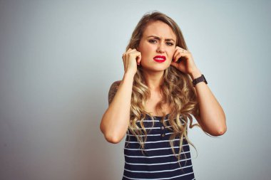Genç güzel kadın yüksek sesle müzik sesi için rahatsız ifade ile parmakları ile kulakları kapsayan beyaz izole arka plan üzerinde duran çizgili t-shirt giyiyor. Sağır kavramı.