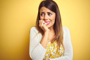 Sarı izole arka plan üzerinde duran ceket giyen genç güzel kadın stresli ve ağız tırnakısırma elleri ile sinirli görünümlü. Anksiyete sorunu.