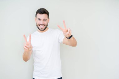 Zafer işareti yapıyor parmak gösterilen kamera seyir gülümseyen izole arka plan üzerinde rahat beyaz t-shirt giyen genç yakışıklı adam. İki numaralı.