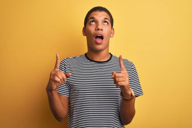 Izole sarı arka plan üzerinde lacivert çizgili t-shirt giyen genç yakışıklı Arap adam şaşırttı ve yukarı bakarak ve parmakları ile işaret ve kolları kaldırdı şaşırttı.