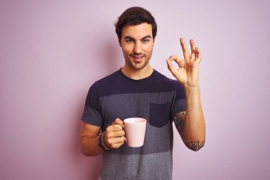 Izole pembe arka plan üzerinde kahve dövme içme fincan ile genç yakışıklı adam parmakları, mükemmel sembolü ile ok işareti yapıyor