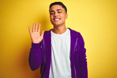 Genç Brezilyalı adam izole sarı arka plan üzerinde duran mor sweatshirt giyen merhaba mutlu ve gülümseyen, samimi hoş geldin jesti söyleyerek Waiving