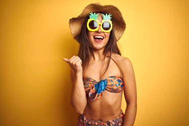 Bikini ve ananas güneş gözlüğü giyen tatilde kadın izole sarı arka plan üzerinde mutlu yüz görünümlü gülümseyerek ve başparmak ile yan işaret.