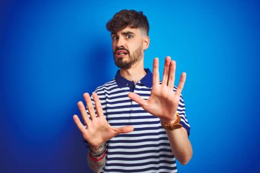 Çizgili polo giyen dövmeli genç adam izole mavi arka plan üzerinde duran korkmuş ve korku ifade ile korkmuş elleriyle jest durdurmak, şok içinde bağırarak. Panik kavramı.
