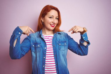 Denim gömlek giyen güzel kızıl saçlı kadın ve izole pembe arka plan üzerinde çizgili t-shirt yüzünde gülümseme ile emin görünümlü, parmakları gururlu ve mutlu kendini işaret.