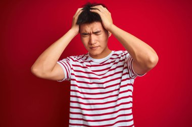 Genç Asyalı Çinli adam çizgili t-shirt giyen izole kırmızı arka plan üzerinde baş ağrısı umutsuz acı ve stresli çünkü ağrı ve migren. Eller baş üstünde.