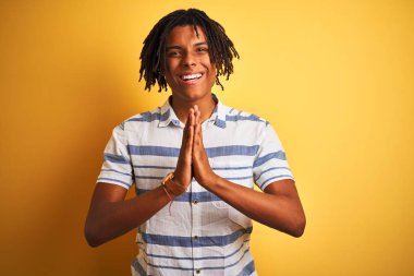 Izole sarı arka plan üzerinde çizgili gömlek giyen dreadlocks ile Afro Amerikan adam birlikte emin gülümseyerek af isteyen elleri ile dua.